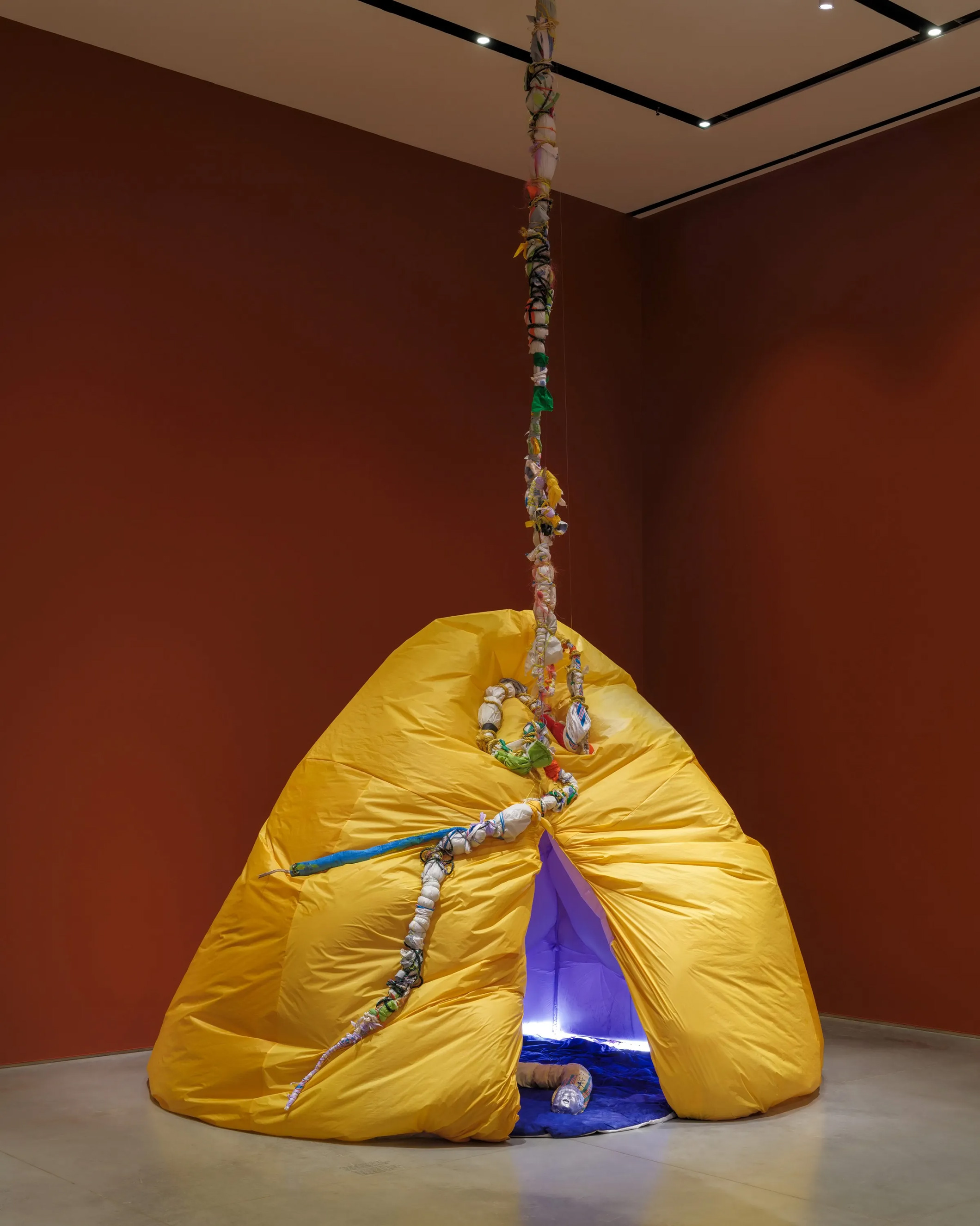 Gallery interior with terracotta-red walls and a gray floor. In the middle is an inflatable yellow hut, split vertically with a blue and purple plush interior. A fabric snake is coiled on the floor.