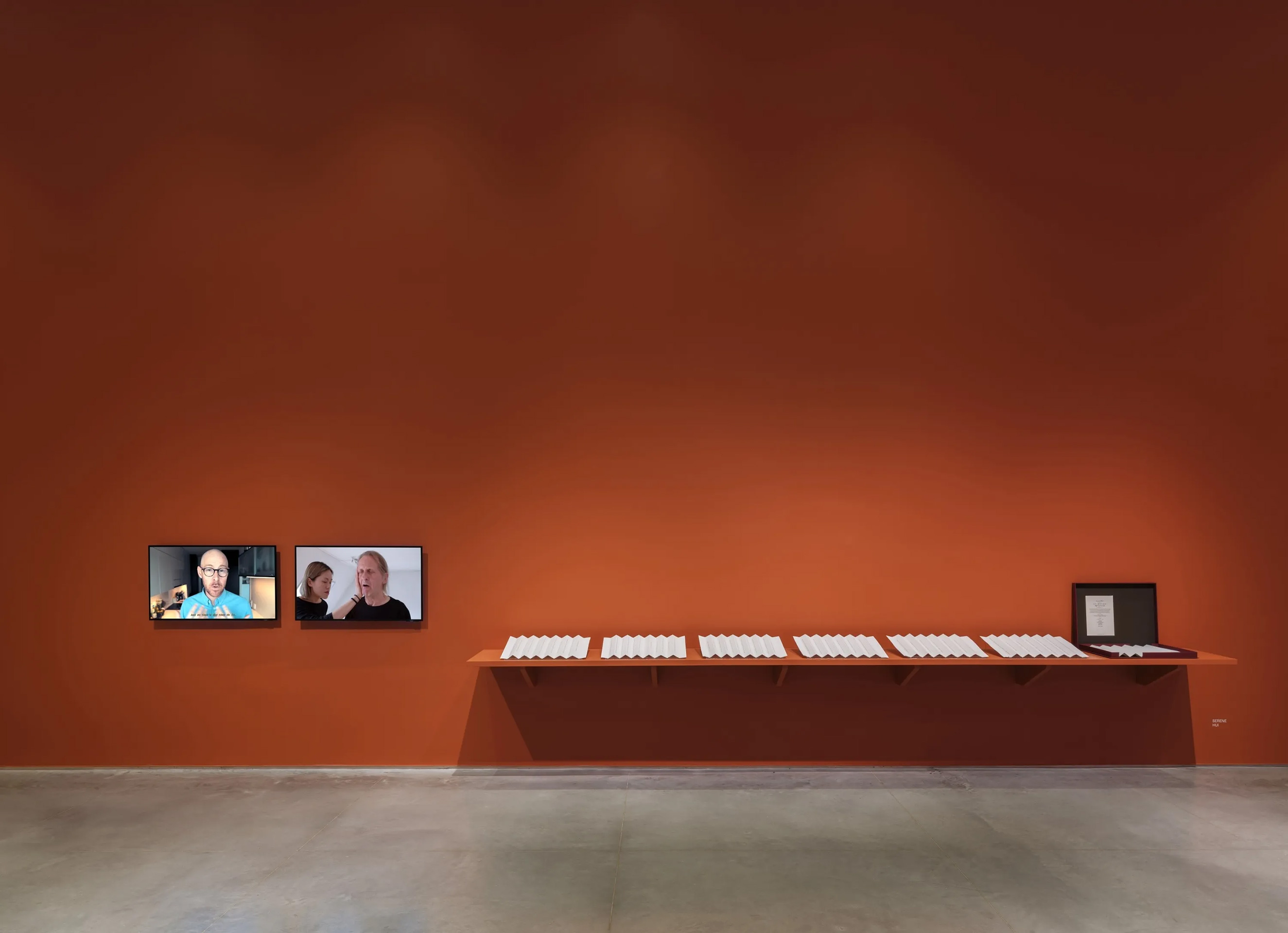 A terracotta-red wall connecting to a gray floor. On the left are two monitors displaying a left and right-channel video. The left-channel video alternates between shots of mouts speaking and footage from popular culture films and television. The right-channel video features a white man and an East Asian woman practicing vocal exercises. To the middle and right, is a terracotta-red shelf holding a row of seven sheets of embossed paper folded accordion-style. The last paper on the end-right is placed in a red box.