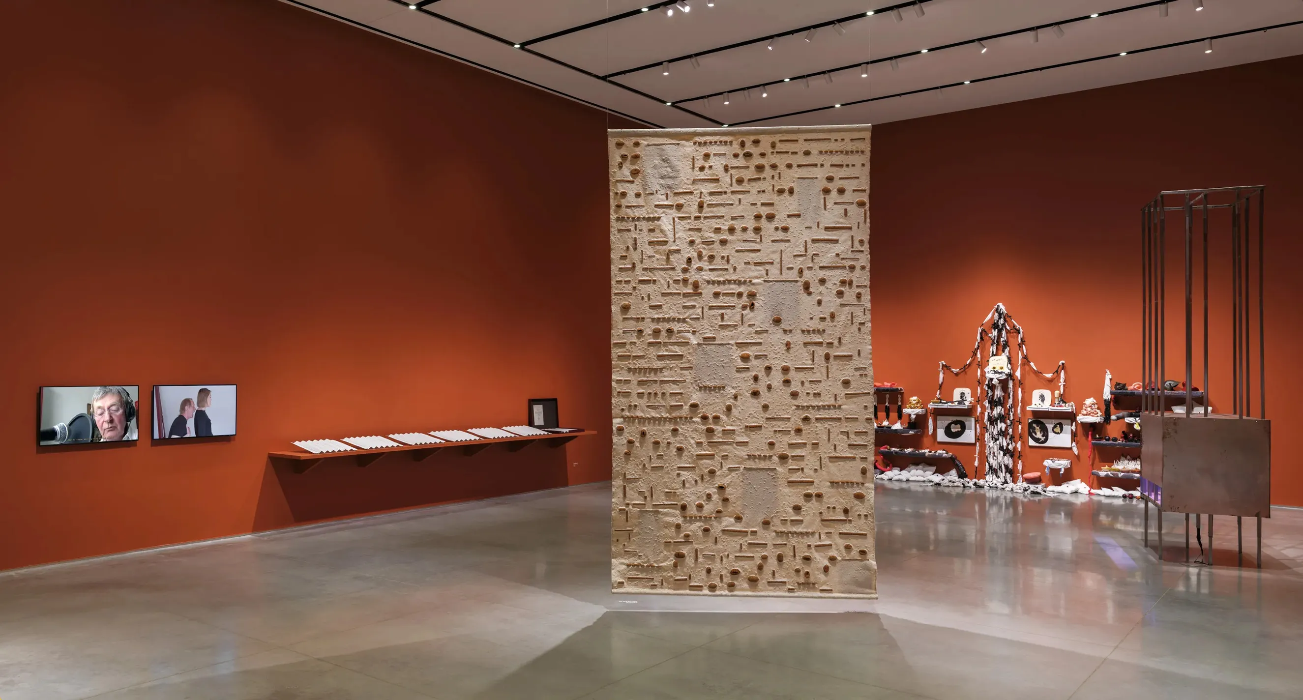 Gallery interior with terracotta-red walls and a gray floor. There are artworks installed on the wall, two tv monitors, a suspended tapestry, and a free-standing cage sculpture.
