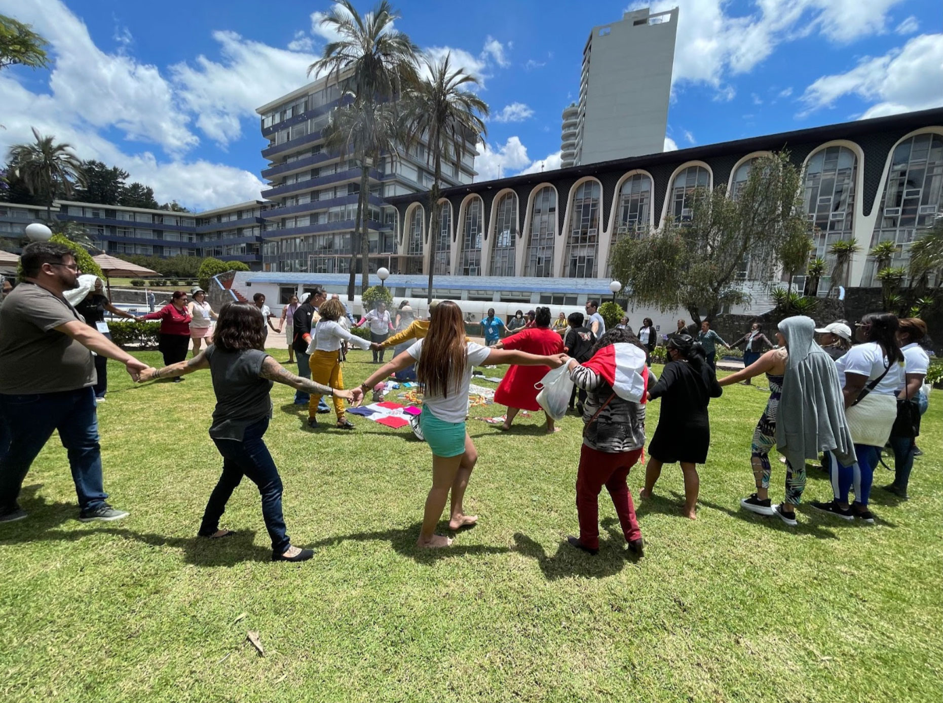 A diverse group of people bonding at an event during the 2022 LUNA retreat in Ecuador stands in a grassy area, holding hands and forming a circle. The scene appears lively and joyful, with participants engaged in an outdoor activity. Buildings and palm trees are in the background under a blue sky with scattered clouds.