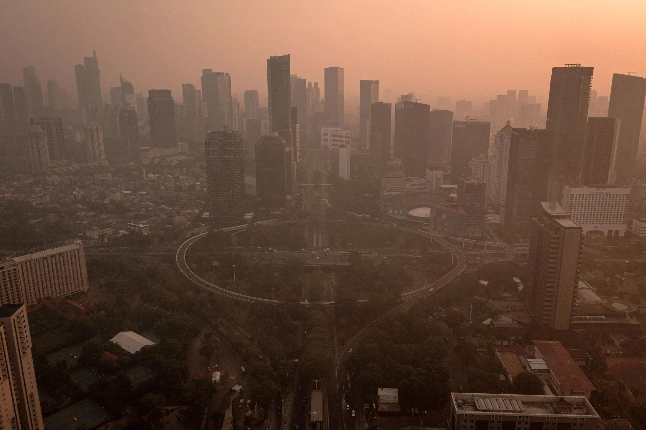 An aerial view of a city skyline at sunset showing numerous high-rise buildings and a large circular road infrastructure in the foreground. The sky is filled with an orange hue, possibly due to haze or smog, casting a warm light over the entire scene.