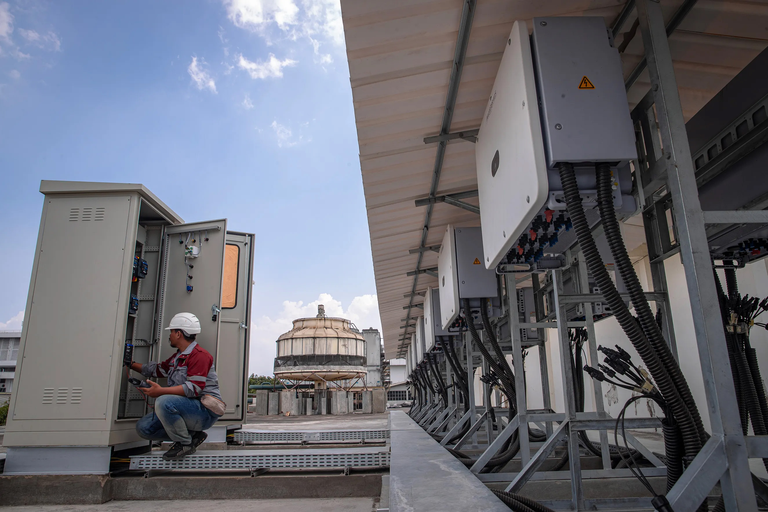 A worker in a hard hat and safety gear squats next to an open electrical panel, inspecting or repairing wiring on a rooftop. Multiple electrical units are lined up beside him, and a large industrial structure is visible in the background under a clear sky.