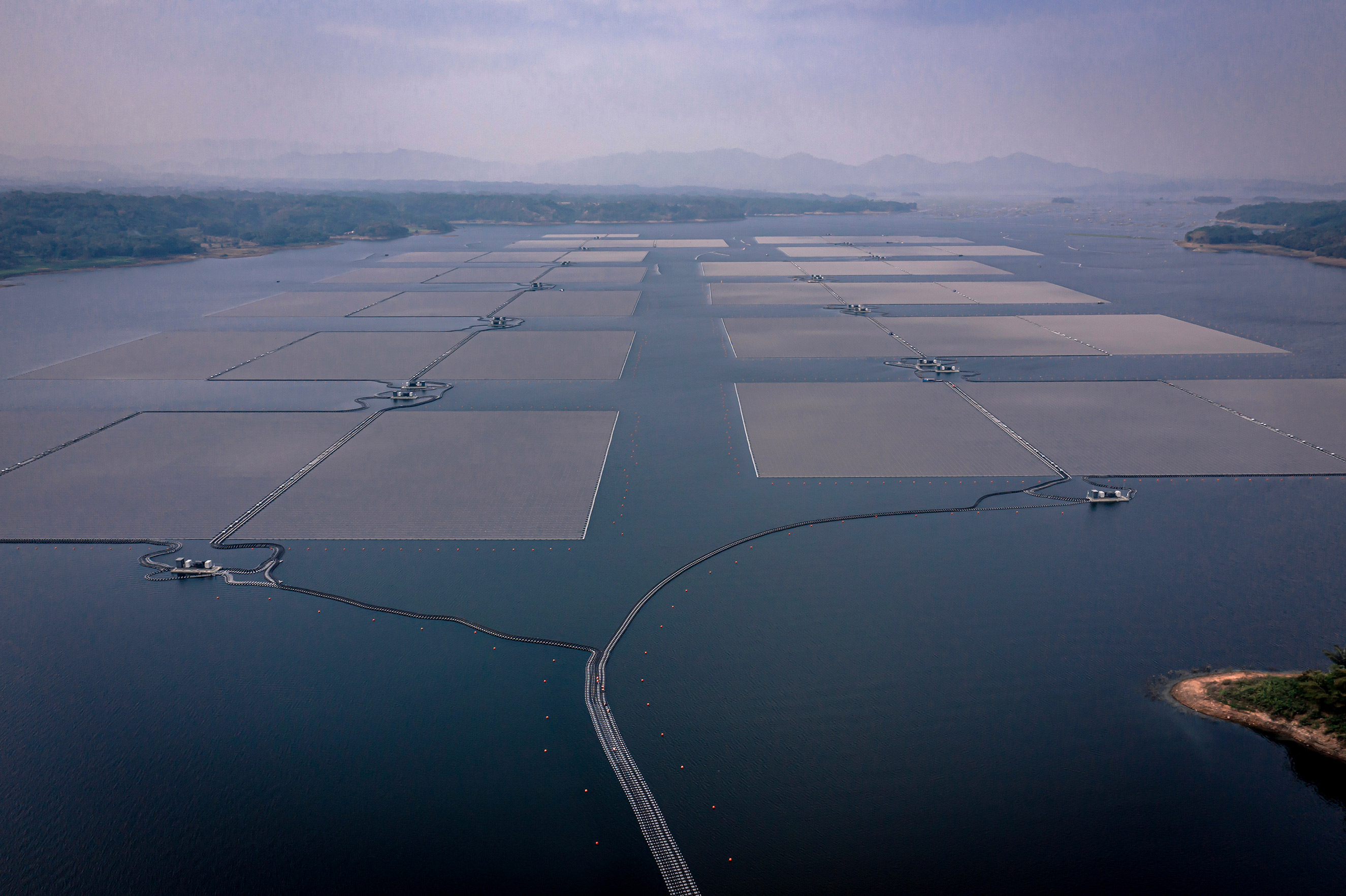 Aerial view of a large floating solar panel farm on a calm body of water, with mist-covered hills and forests in the background. The grid-like arrangement of solar panels stretches across the water, interconnected by walkways and cables.