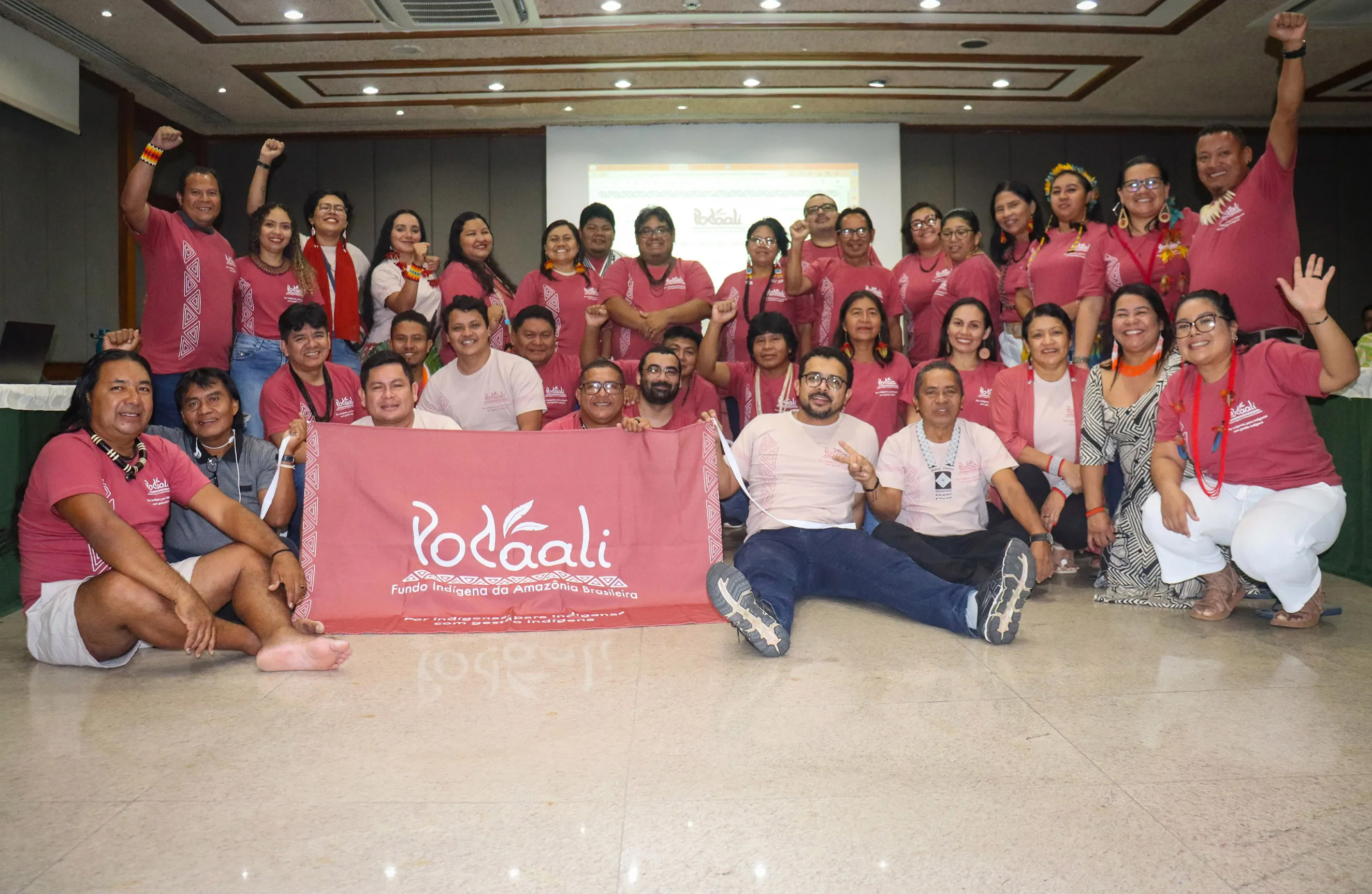 Members of Podáali posing together in a conference room, wearing matching pink T-shirts with the word "Podáali" on them. They are smiling and some are making celebratory gestures. The group holds a banner with the "Podáali" text, and a screen is visible in the background.