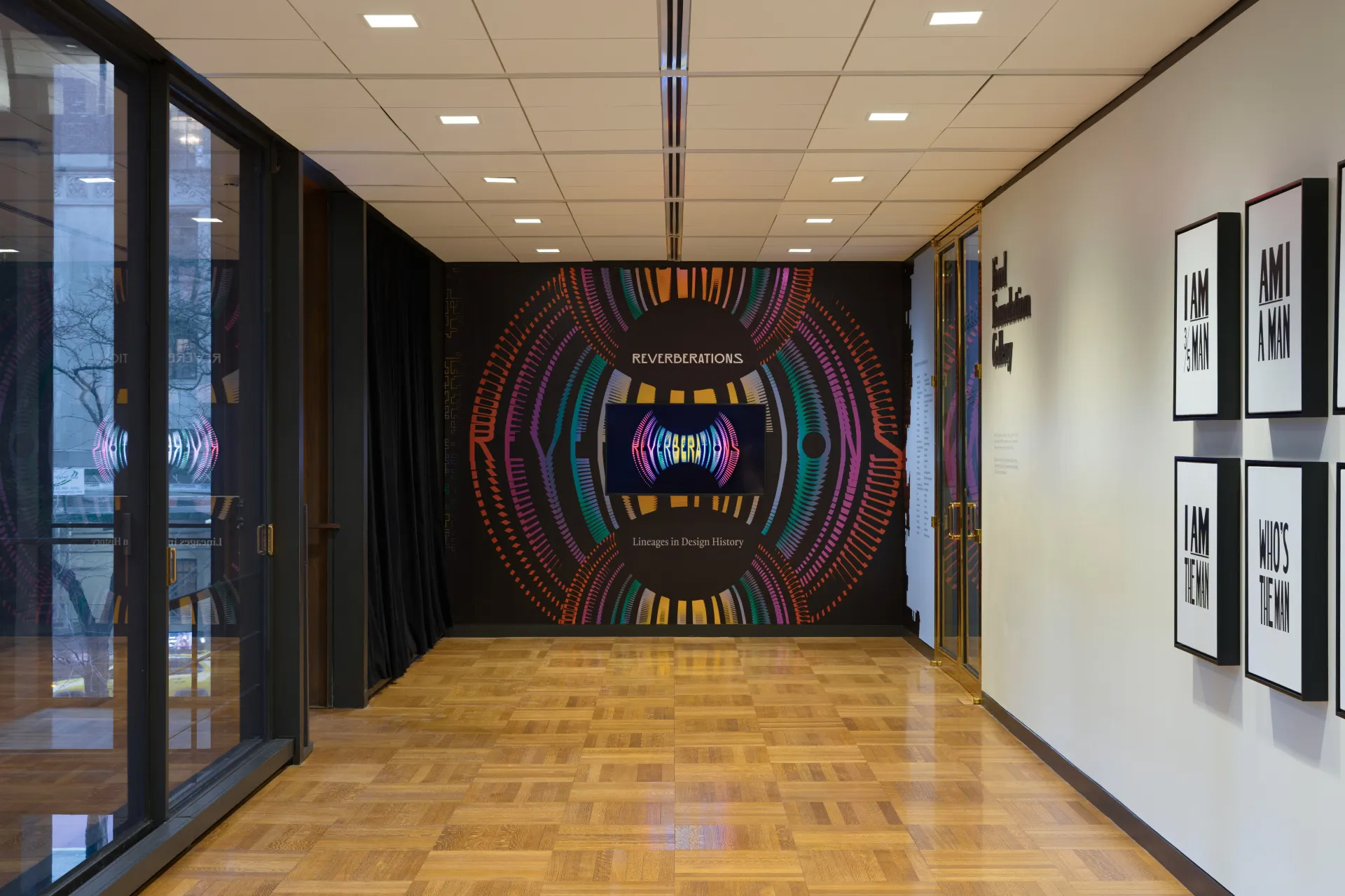 A hallway with wood flooring and framed text art on the right wall. The end wall features a vibrant, circular abstract pattern with the text REVERBERATIONS with a digital screen in the middle displaying the same animated text.