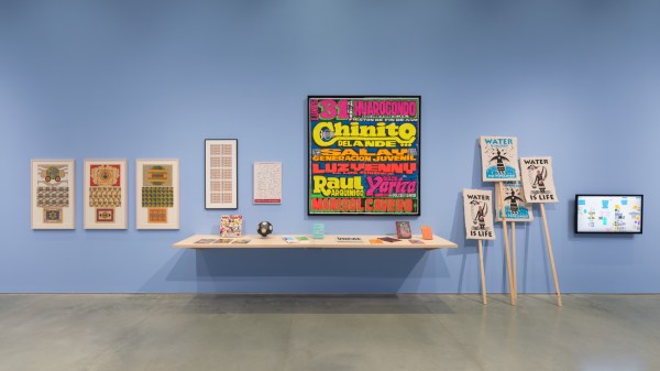 An exhibition wall displays colorful posters, framed artworks, and smaller items and publications on a shelf. The centerpiece is a vibrant, text-heavy poster. To the right, posters on wooden sticks say “Water is Life”.