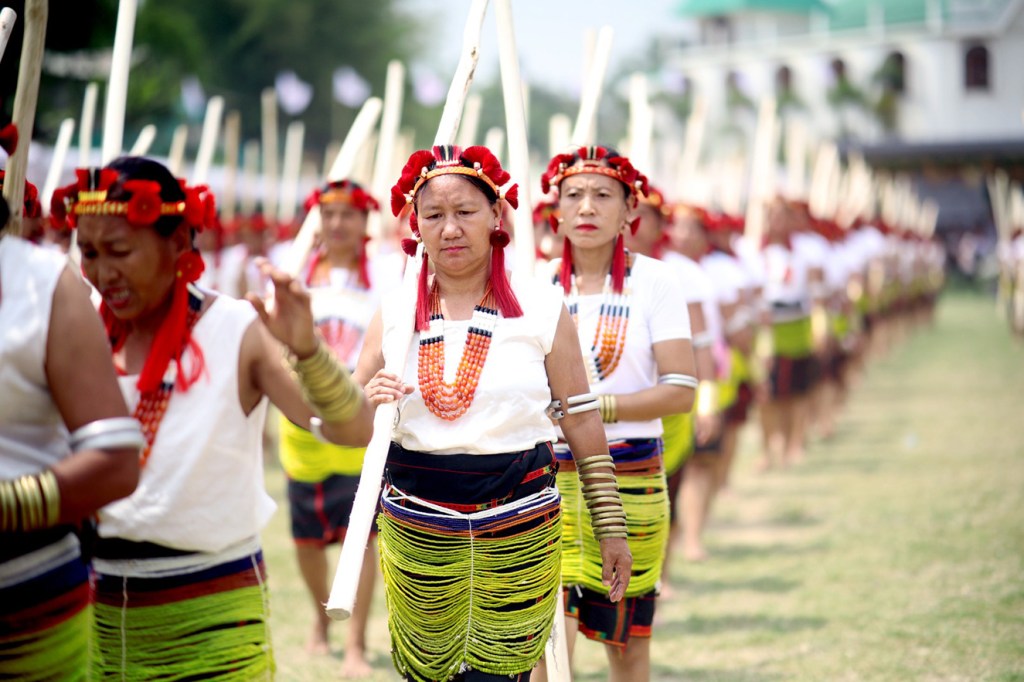 A group of women in traditional attire, decorated with colorful beads and headdresses, are marching in formation holding sticks. The scene appears outdoors in a grassy area with a building and trees in the background.