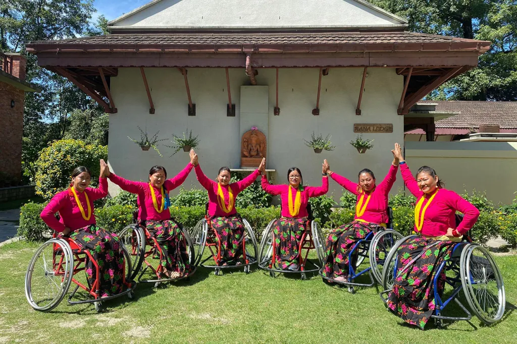 Six women in wheelchairs wearing matching red tops and floral skirts perform a synchronized dance pose outdoors, in front of a building. Their arms are raised, and they wear yellow necklaces, with greenery in the background.