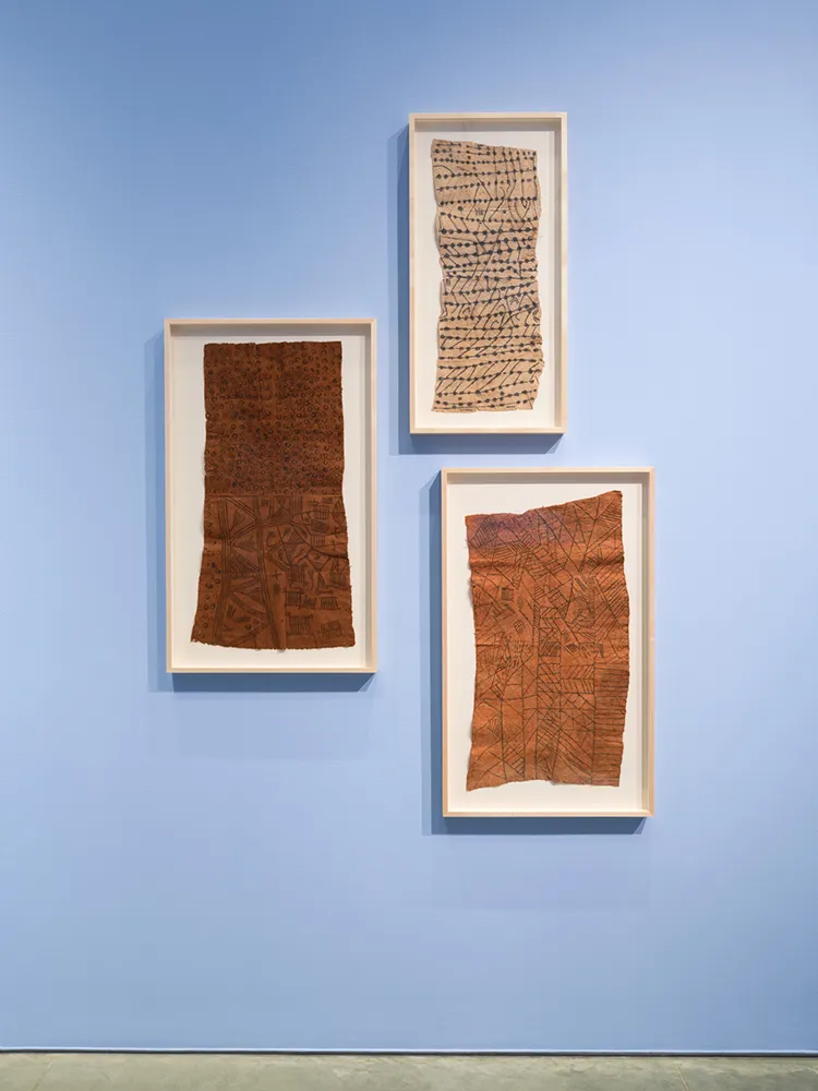 Three rectangular framed paintings on tree bark on sky blue gallery walls.