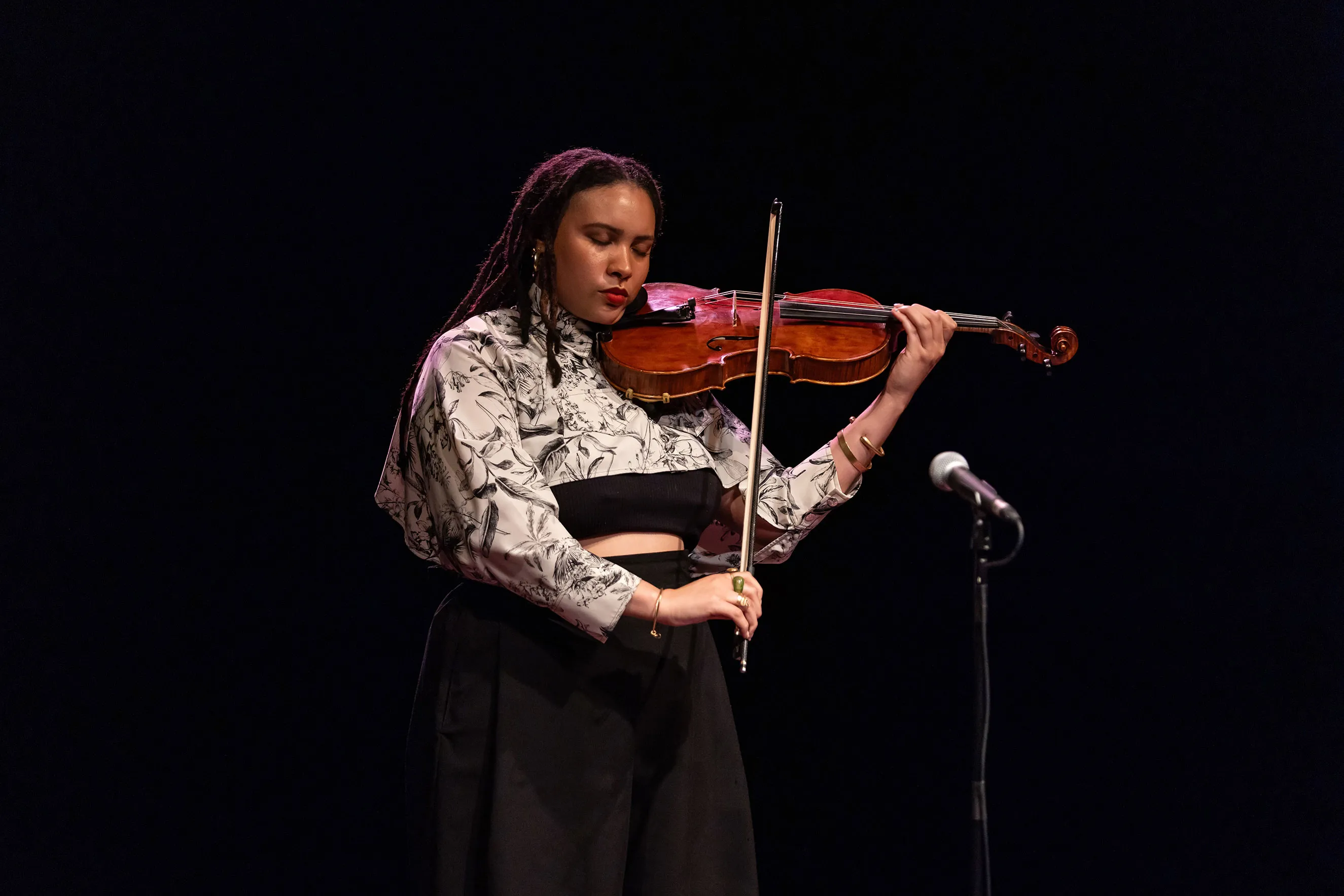 A violinist wearing black and white standing at a microphone and playing the violin.