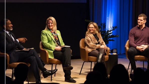 A panel of four people sit on stage during the Ideas At Ford event. Three are smiling as one speaks. They are seated in chairs with a blue-lit backdrop behind them. Left to right they are Charles Blow, Liz Shuler, Jessica Berman, and Carl Nassib.