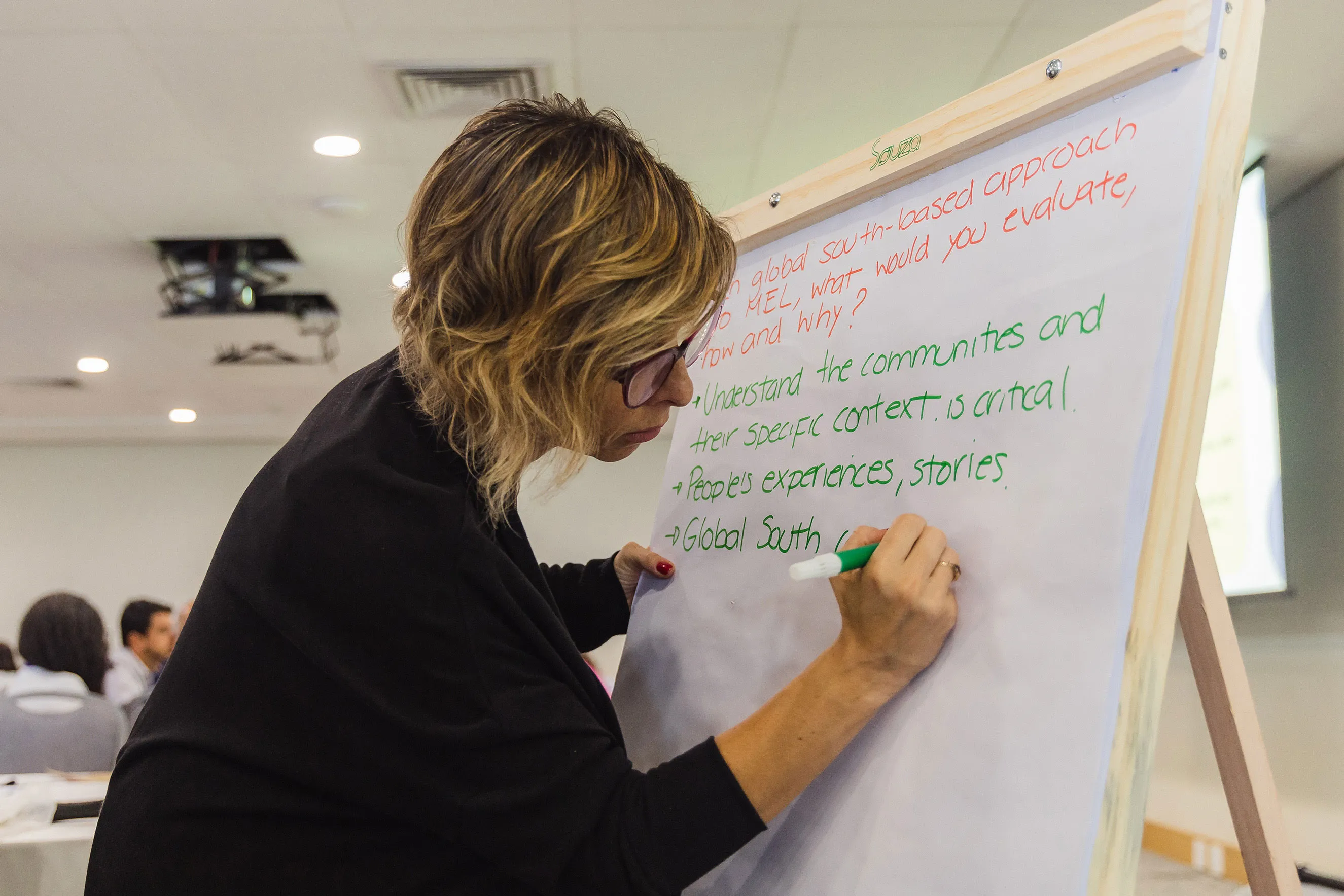 A woman with glasses writes on a large flip chart with a green marker in a classroom or meeting room. The chart has bullet points about understanding communities and the Global South. Other people are visible in the background.