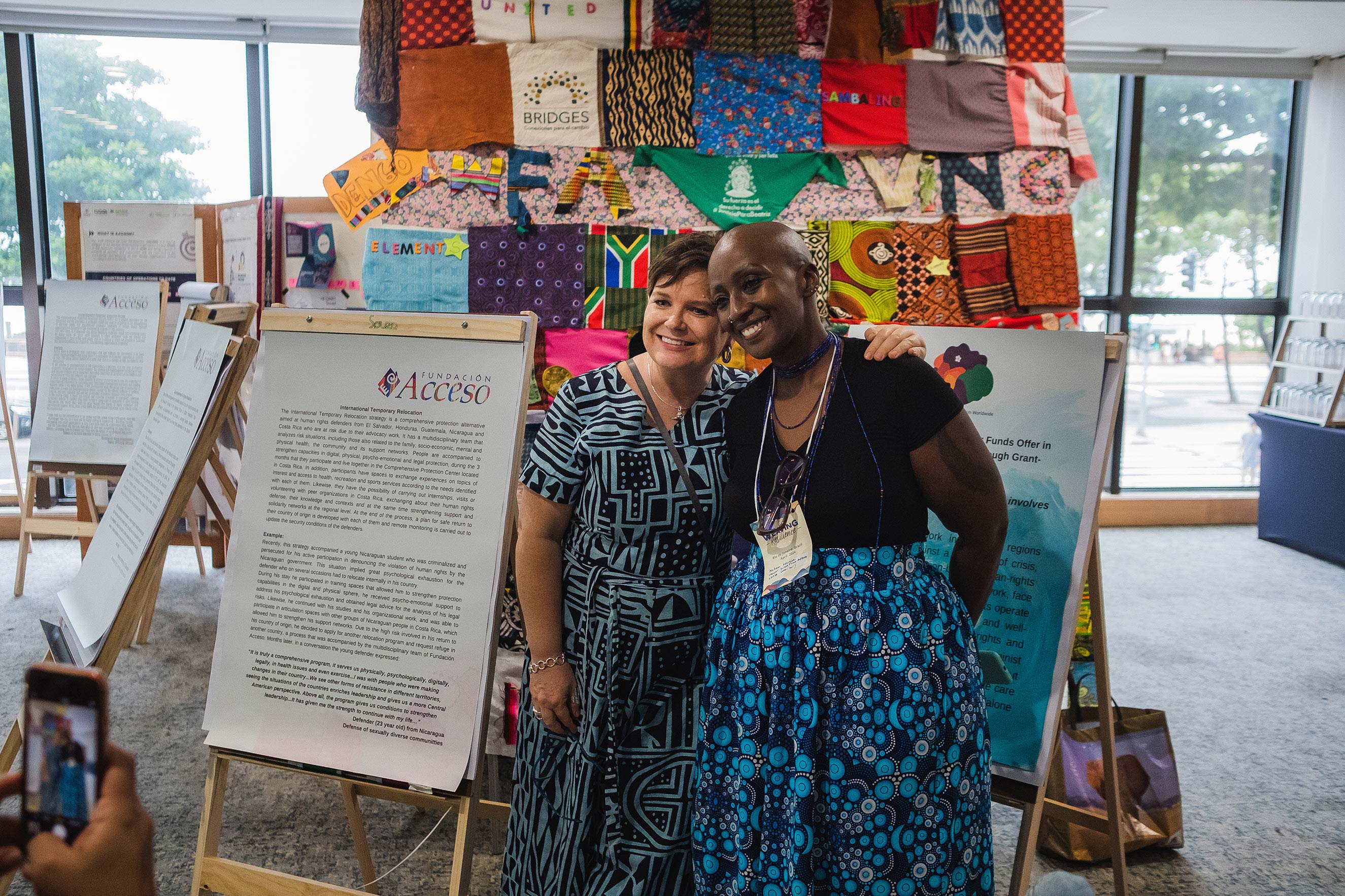 Two women smile and pose together in front of colorful fabric displays and informational boards at an indoor event, while someone takes their photo. Large windows in the background let in natural light.