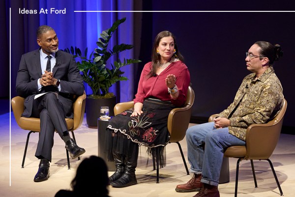 Three people sit on stage in a panel discussion. The man on the left, Charles Blow, wears a suit. The woman, Aimee Roberson, in the middle, gestures while speaking. The man on the right, Tristan Ahtone, wears glasses and a patterned shirt. A plant is in the background.