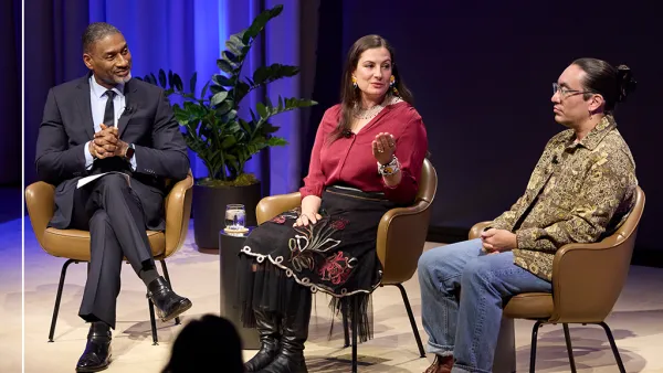 Three people sit on stage in a panel discussion. The man on the left, Charles Blow, wears a suit. The woman, Aimee Roberson, in the middle, gestures while speaking. The man on the right, Tristan Ahtone, wears glasses and a patterned shirt. A plant is in the background.