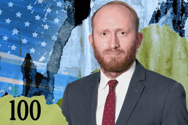 A man with short red hair and a beard, wearing a gray suit, white shirt, and red polka dot tie stands in front of an abstract background with stars, blue, black, and green paint, and the number 100 in the lower left corner.