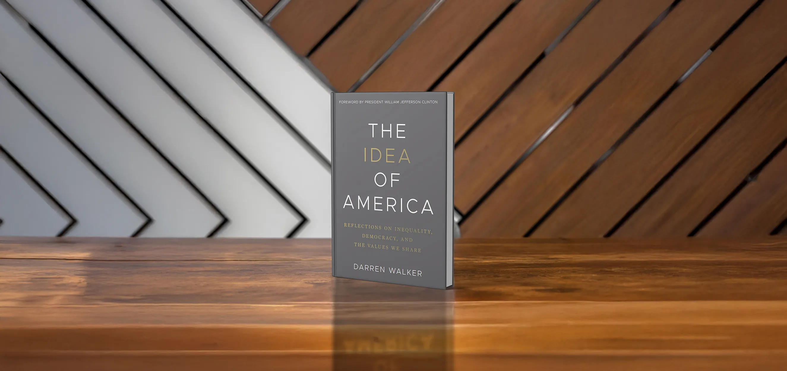 A book titled The Idea of America by Darren Walker stands upright on a wooden surface with a diagonally patterned wooden wall in the background.