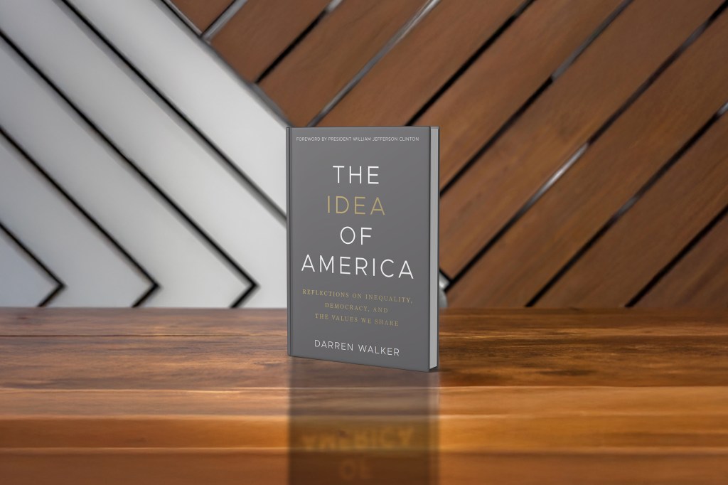 A book titled The Idea of America by Darren Walker stands upright on a wooden surface with a diagonally patterned wooden wall in the background.