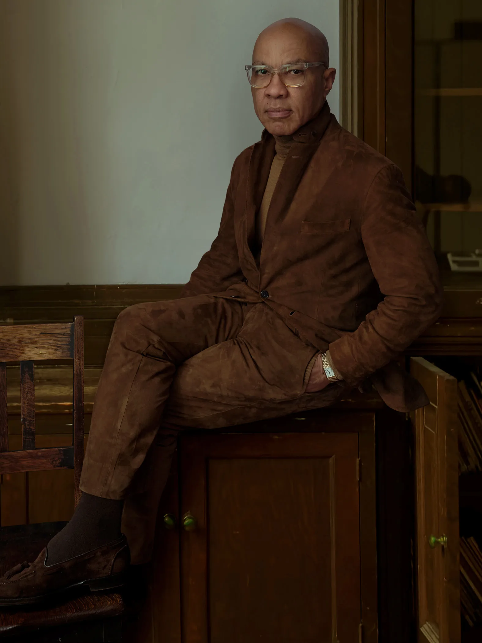 A formal portrait of Darren Walker, a bald man wearing glasses and a brown suede suit. He is seated on top of a dark wooden cabinet, looking directly at the camera with a serious expression.