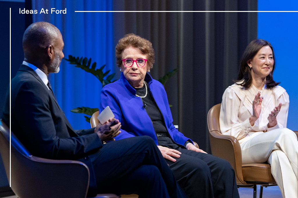 Three people are sitting in conversation against a blue curtain background. The group includes, from left to right, Charles Blow in a dark suit, Billie Jean King in black with a blue blazer, and Clara Wu Tsai in a beige white and trousers.