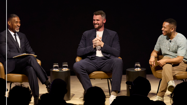 Three people are sitting in conversation against a dark curtain background. The group includes, from left to right, Charles Blow in a black suit, white shirt, and black tie. Kevin Love in a dark blue suit and white t-shirt, and Yolo Akili in a knitted top and khaki trousers.