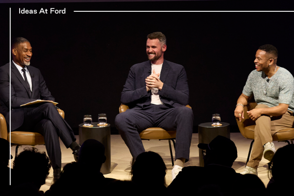 Three people are sitting in conversation against a dark curtain background. The group includes, from left to right, Charles Blow in a black suit, white shirt, and black tie. Kevin Love in a dark blue suit and white t-shirt, and Yolo Akili in a knitted top and khaki trousers.