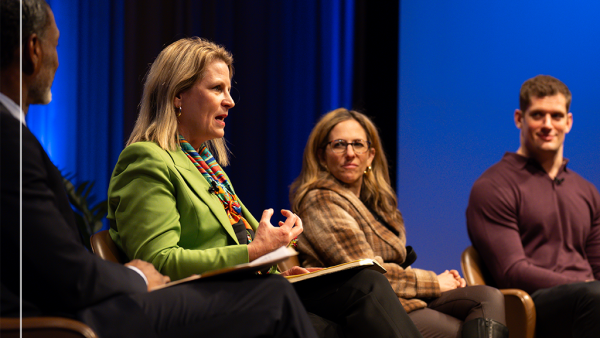 A panel of four people sit on stage during the Ideas At Ford event. Three are smiling as one speaks. They are seated in chairs with a blue-lit backdrop behind them. Left to right they are Charles Blow, Liz Shuler, Jessica Berman, and Carl Nassib.