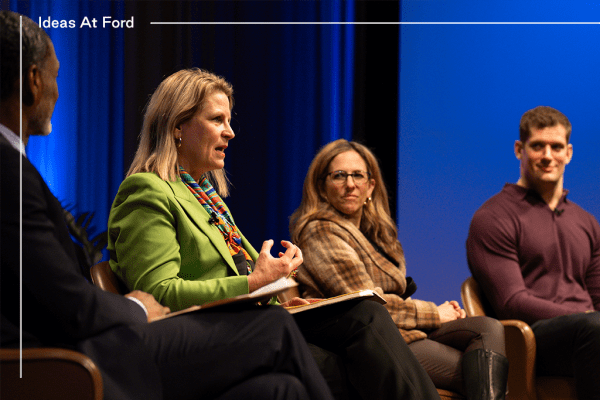 A panel of four people sit on stage during the Ideas At Ford event. Three are smiling as one speaks. They are seated in chairs with a blue-lit backdrop behind them. Left to right they are Charles Blow, Liz Shuler, Jessica Berman, and Carl Nassib.