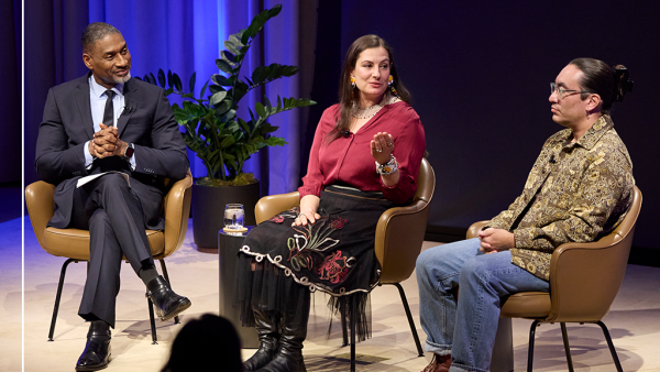 Three people sit on stage in a panel discussion. The man on the left, Charles Blow, wears a suit. The woman, Aimee Roberson, in the middle, gestures while speaking. The man on the right, Tristan Ahtone, wears glasses and a patterned shirt. A plant is in the background.