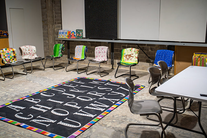 A classroom scene features several chairs with colorful, patterned fabric backs, an alphabet rug, and a table with more chairs. Books are visible on a ledge above the chairs, and whiteboards are in the background.