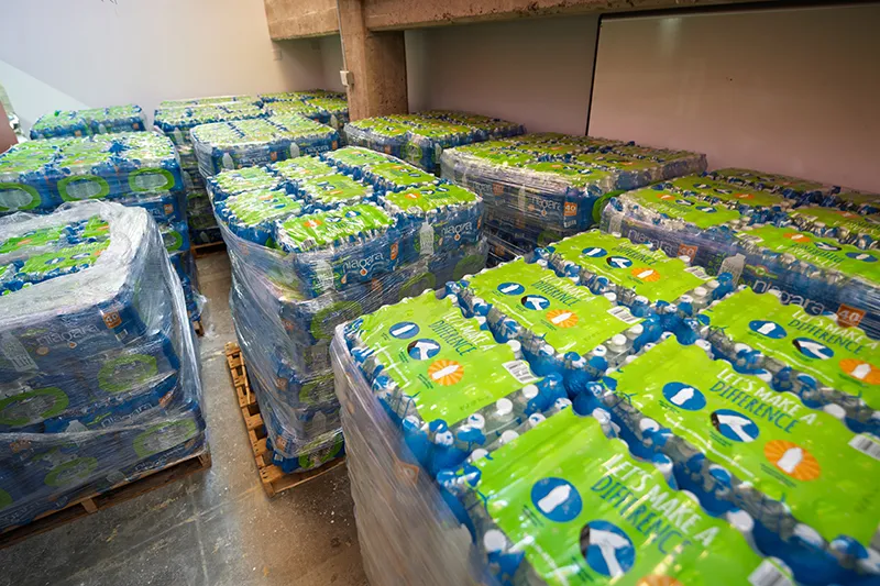A room is filled with pallets stacked high with cases of bottled water, each bottle featuring a green label with the text "LET'S MAKE A DIFFERENCE." The bottles are clear, showing the water inside.