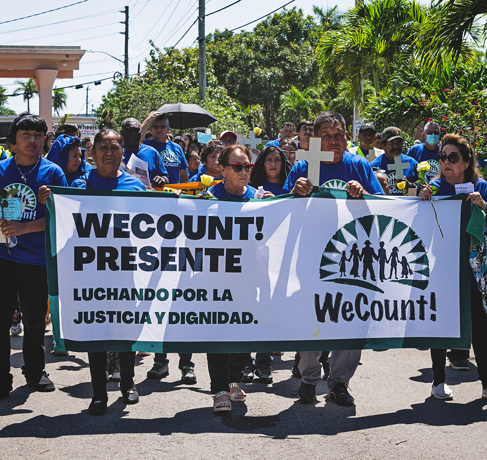 A diverse group of people marches outdoors under a clear sky, holding a large banner that reads "WECOUNT! PRESENTE LUCHANDO POR LA JUSTICIA Y DIGNIDAD" with a graphic of silhouetted figures and "WeCount!".