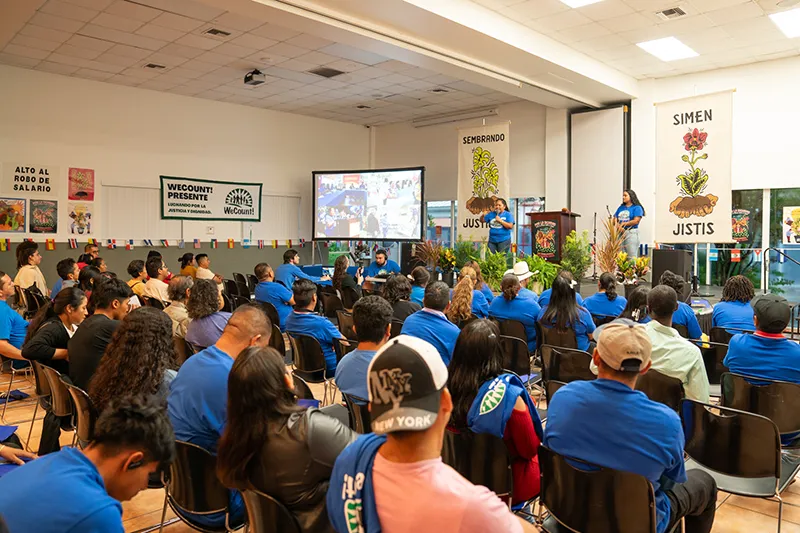 A large group of people wearing blue shirts are seated, facing a stage with a large screen displaying an image. Two people are speaking at podiums, and banners hang in the background with text and plant illustrations.