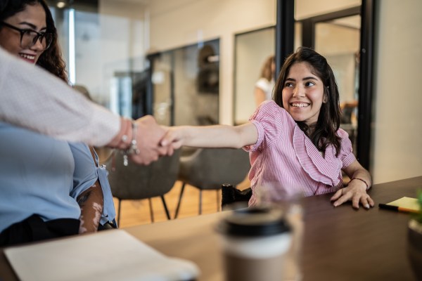 A woman sitting at a table extends her arm to shake hands with another person across from her in an office meeting room.