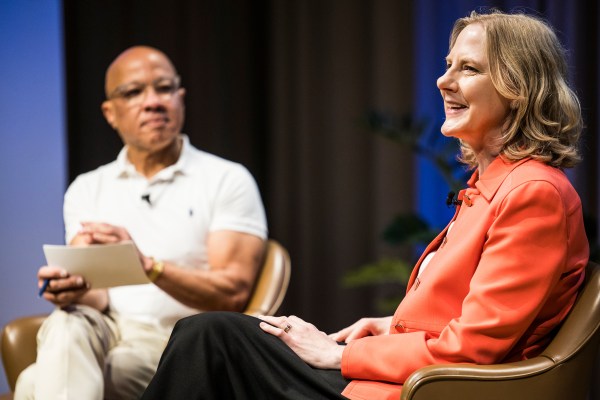 A candid photo of **Heather Gerken in a bright orange blazer** smiling while seated on the right. In the blurred background on the left, **Darren Walker** sits in a white polo shirt, holding papers.