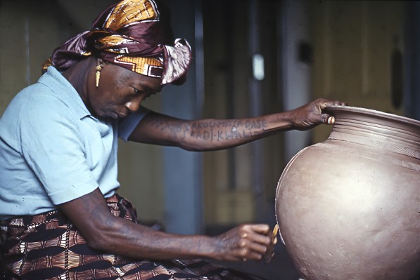 A Black woman carving a geometric design into a clay pot. Her arms are heavily tattooed and she’s in deep focus.