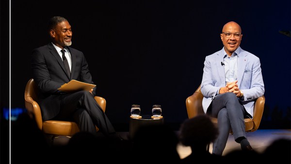 Two people are sitting in conversation against a dark curtain background. The group includes, from left to right, Charles Blow in a black suit, white shirt, and black tie, and Darren Walker wearing a light blue blazer, striped collar shirt and dark blue slacks.