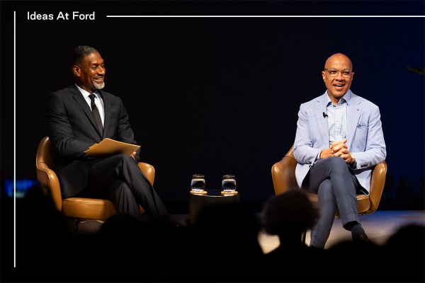 Two people are sitting in conversation against a dark curtain background. The group includes, from left to right, Charles Blow in a black suit, white shirt, and black tie, and Darren Walker wearing a light blue blazer, striped collar shirt and dark blue slacks.