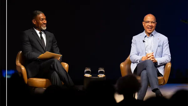 Two people are sitting in conversation against a dark curtain background. The group includes, from left to right, Charles Blow in a black suit, white shirt, and black tie, and Darren Walker wearing a light blue blazer, striped collar shirt and dark blue slacks.