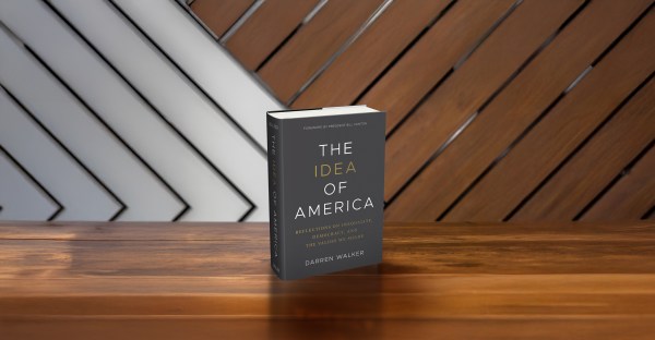A book titled The Idea of America by Darren Walker stands upright on a wooden surface with a diagonally patterned wooden wall in the background.