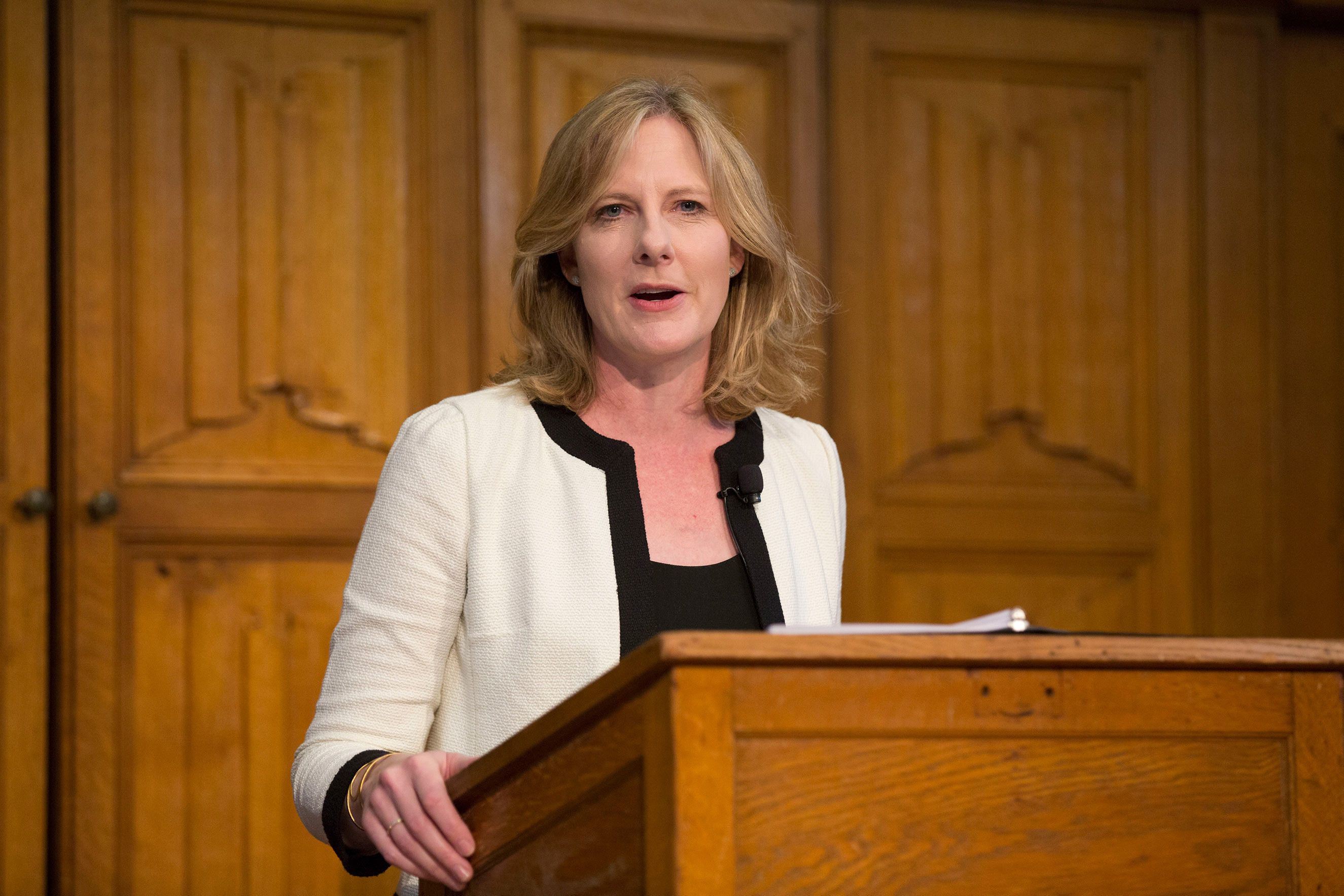 Heather Gerken, the 11th President of the Ford Foundation, wearing a white jacket with black trim, stands at a wooden podium, speaking in front of wooden panel doors.