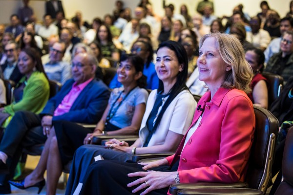 A Heather Gerken in a bright pink jacket** sits in the front row of an auditorium, smiling and looking forward. She is seated next to three other people, with a large, diverse crowd blurred in the rows behind them.