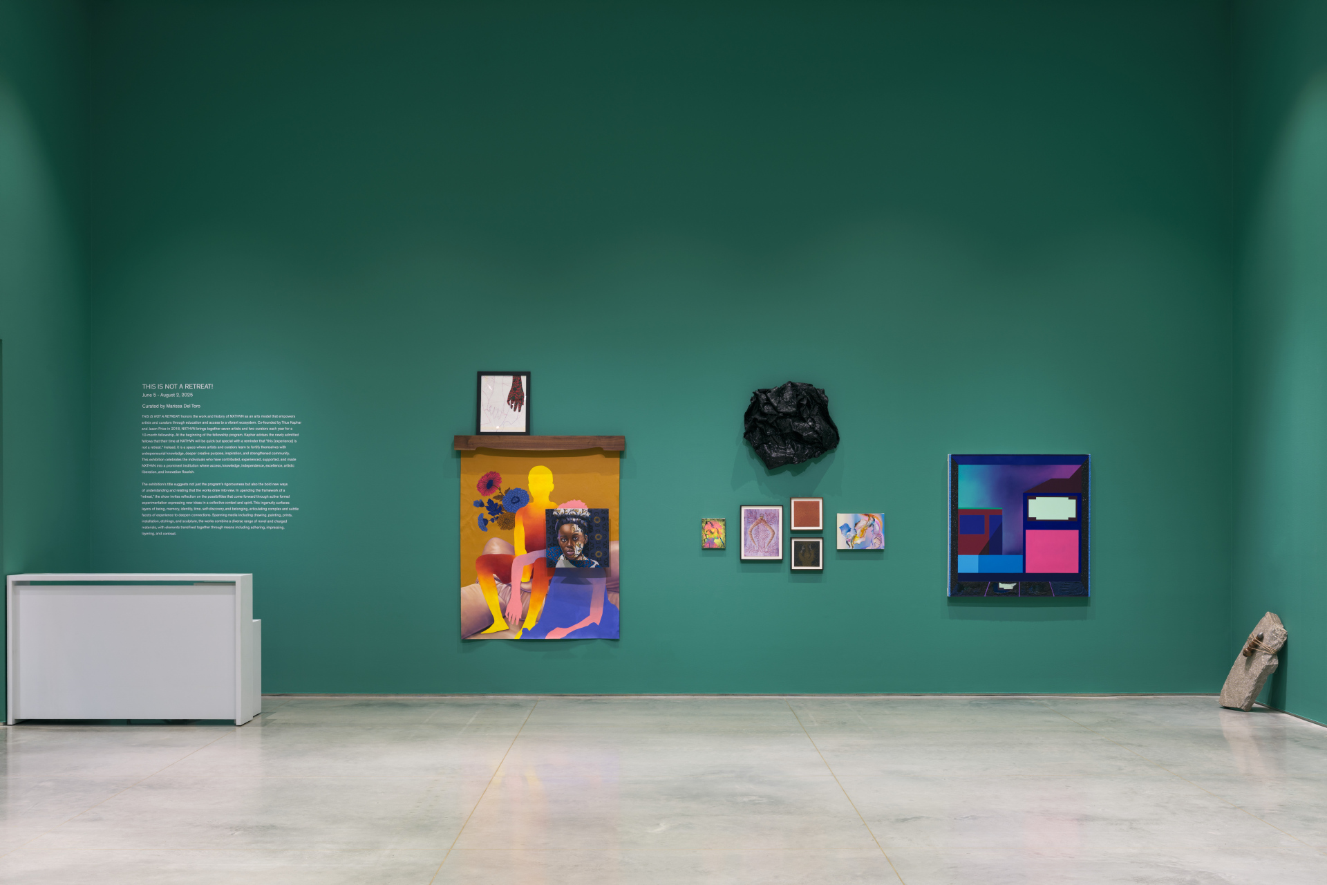 A green gallery interior with a white desk to the left, various hanging artworks of different mediums and a stone sculpture.