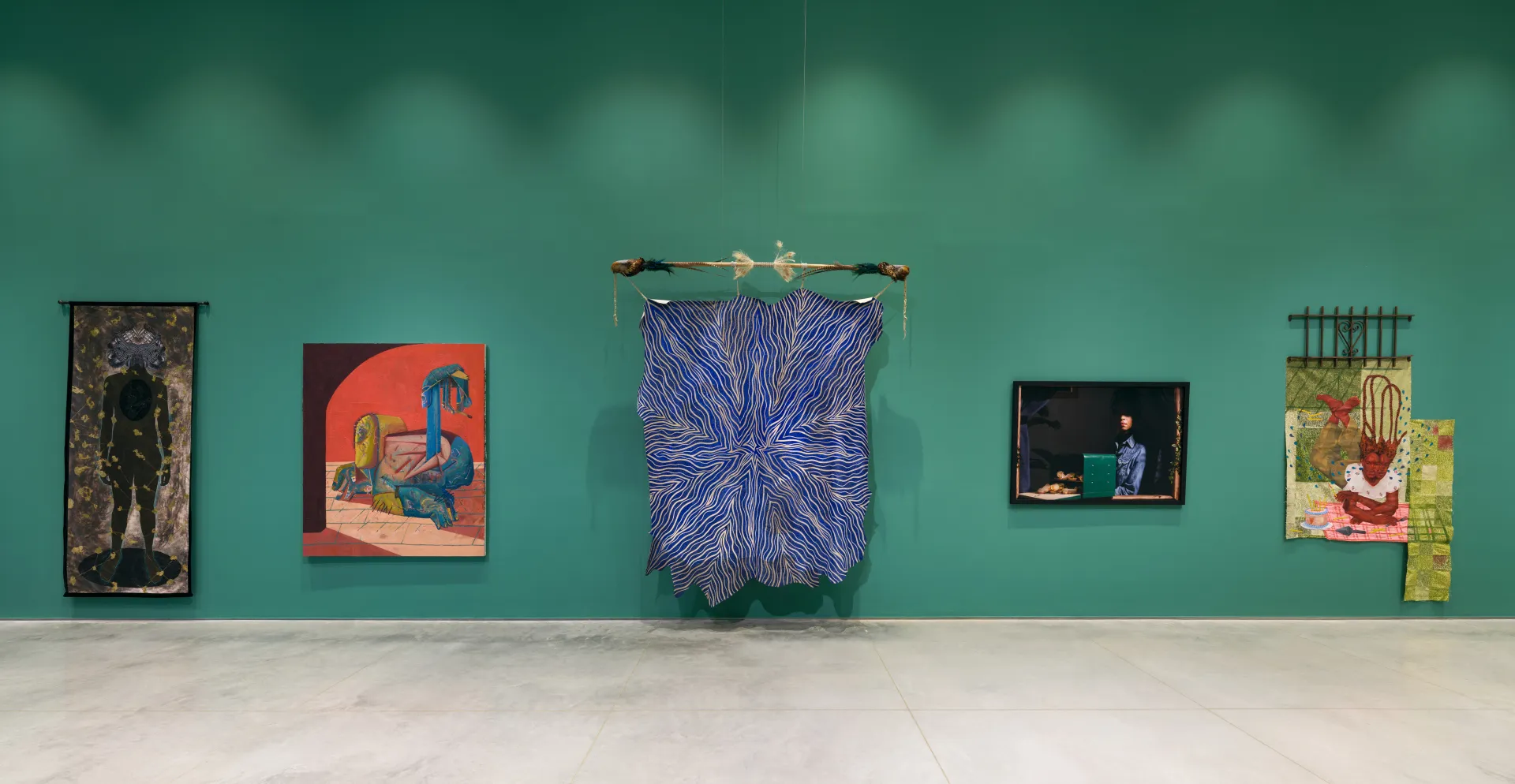 A green gallery wall with various hanging artworks that are multi-media, painted canvas, and photography.