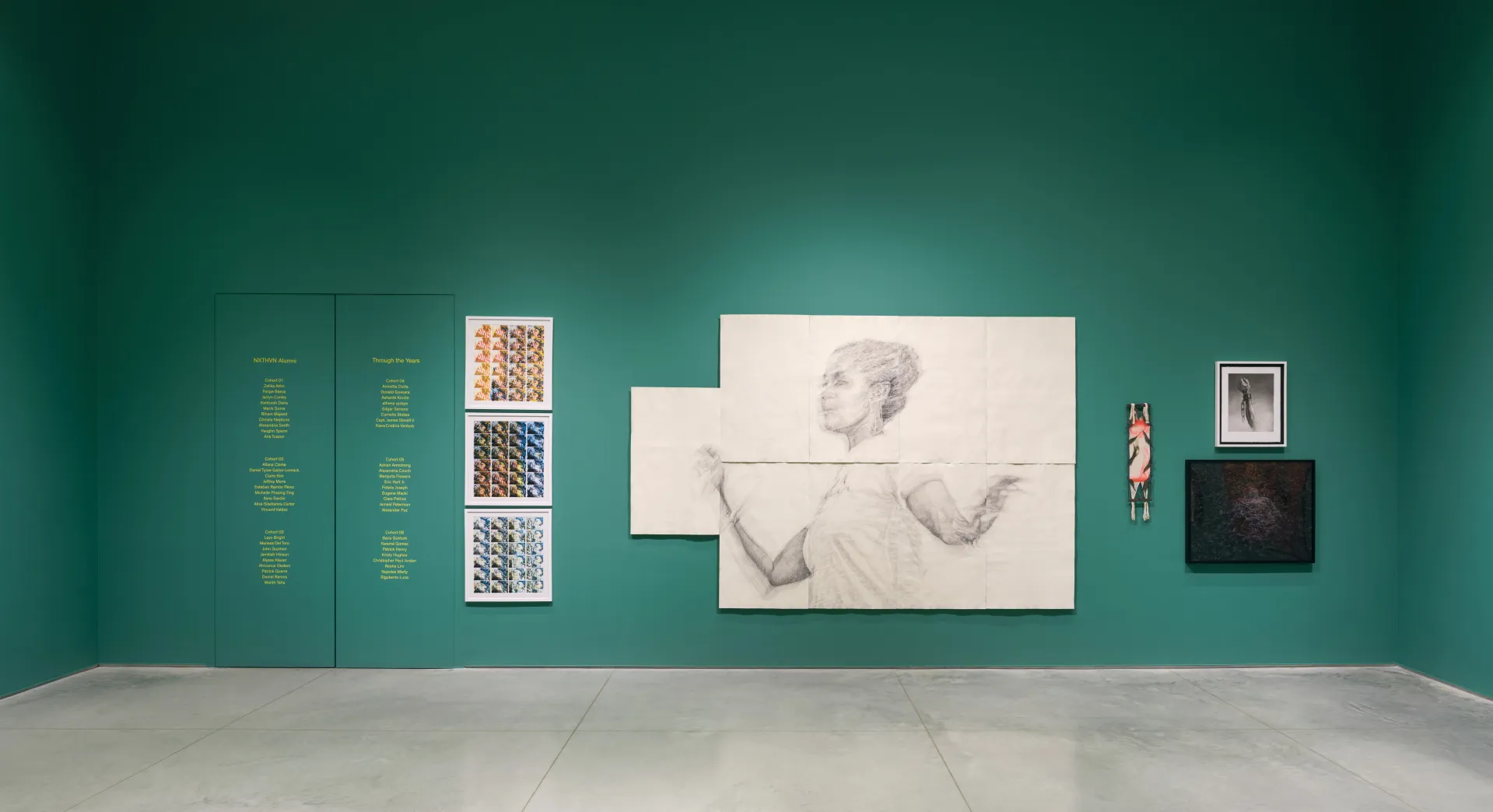 A green gallery wall with different artworks, one of the works features a graphite drawing of a Black woman on sheets of paper.