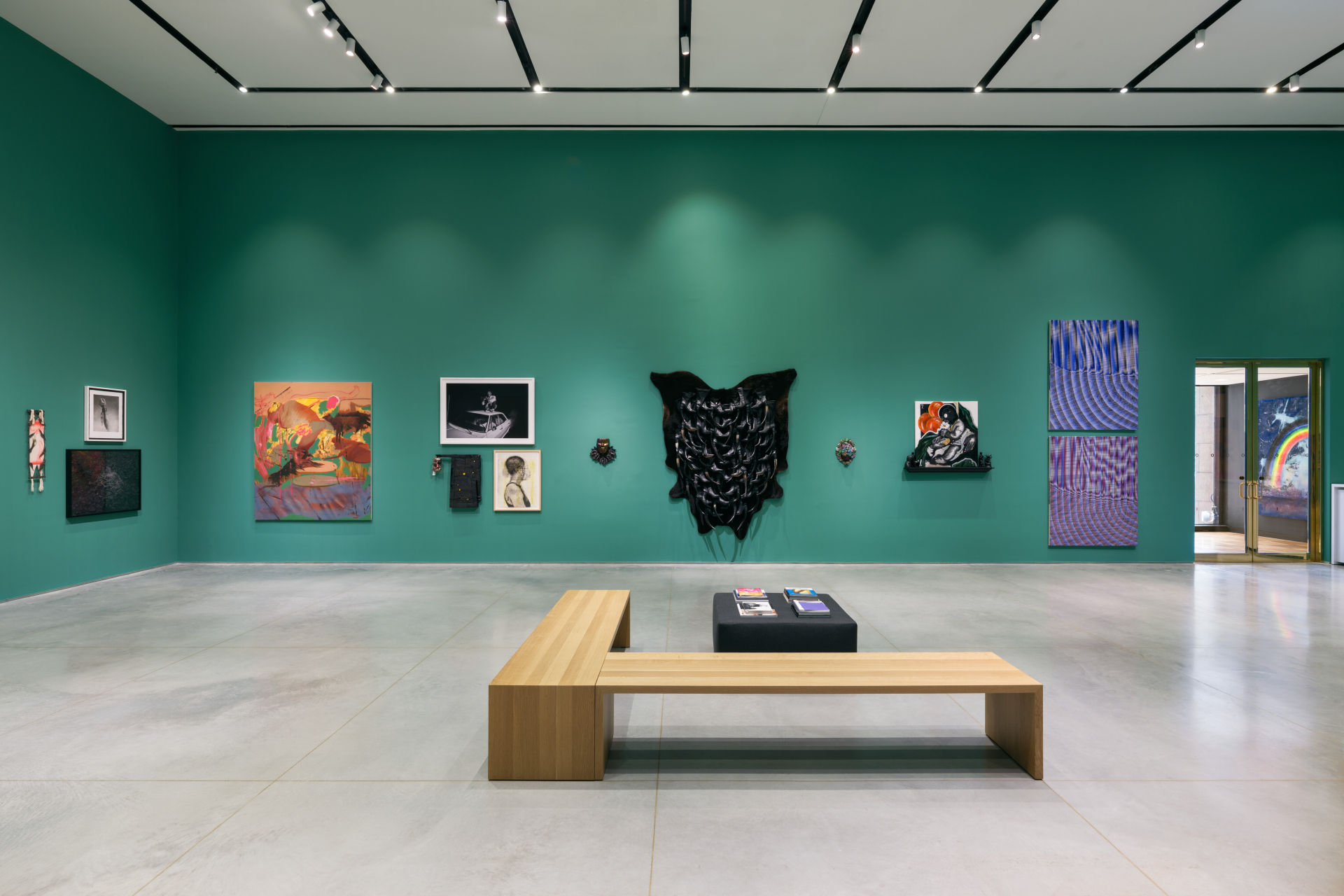 An installation view of a gallery exhibition featuring a salon-style wall of works. There is an L-shaped bench and cushions for guests to sit and read.