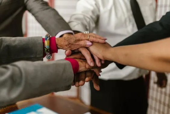 A close-up of four hands, belonging to a diverse group of people, stacked on top of each other in a gesture of unity and teamwork. One hand has colorful bracelets and wrinkled skin.