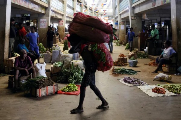 A person walks through a busy, covered market carrying a very large, heavy sack of produce on their back. People and various vegetables, including onions and eggplants, are visible in the stalls and aisles around them.