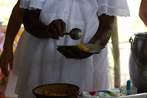 A close-up of a person in a white top with eyelet lace, holding a green leaf. They use a ladle to place a yellow rice onto the leaf, as part of a meal preparation.