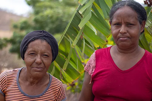 A headshot of two women standing outdoors. The woman on the left wears a black head wrap and a striped shirt, and the woman on the right wears a pink shirt. They are looking directly at the camera.